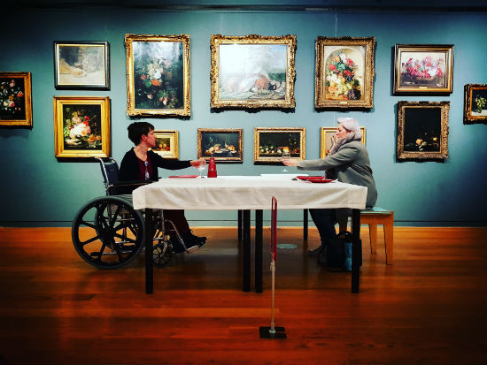 A woman sits across a table from a man in a wheelchair. They are raising a glass to each other. Behind them is a wall covered in paintings.