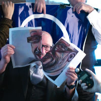 A seated man rips apart a painting of himself revealing his face. Behind him stands a woman and man holding up a blue banner. In front of him a man.