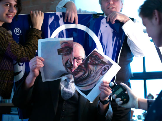 A seated man rips apart a painting of himself revealing his face. Behind him stands a woman and man holding up a blue banner. In front of him a man.