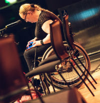 A woman in a wheelchair works on a tablet. In front of her is a microphone and to the left are music stands.