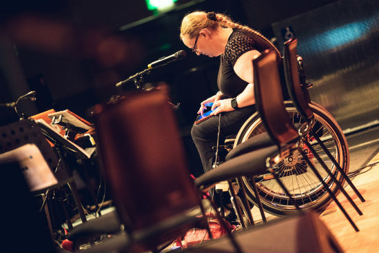 A woman in a wheelchair works on a tablet. In front of her is a microphone and to the left are music stands.