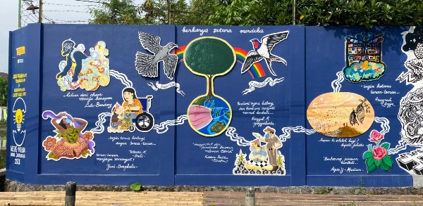 Outdoor mural on a blue wall showing a variety of annotated illustrations of people, nature and places, all connected to one another by wavy lines. At the centre is a globe from which a giant tree grows, flanked by birds on each side with a rainbow between them.