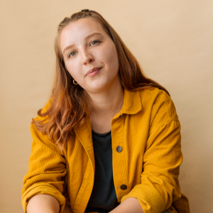 A white woman in her early 20s with mid length brown hair and bluey-green eyes. She wears a black top with an orange overshirt.