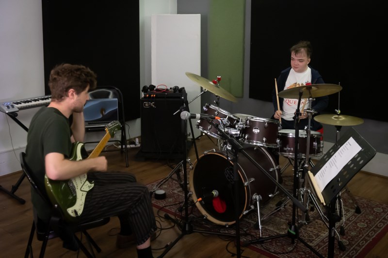 Two musicians, one a teenager playing the drums and other a man in his thirties playing the guitar in a rehearsal studio