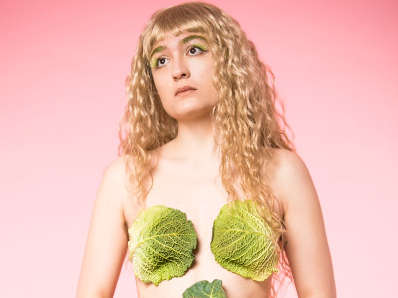 A slim white femme person with wavy blonde hair, naked except for two pieces of lettuce covering her chest area