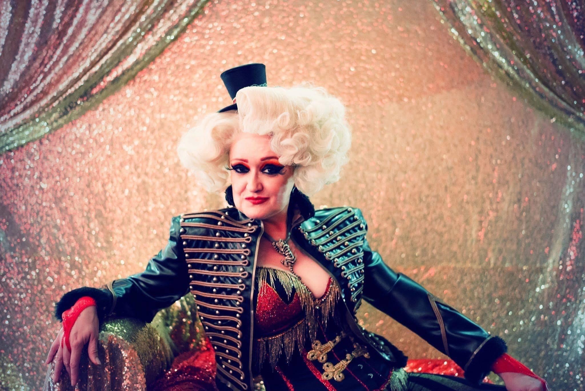 Cabaret performer Diva Hollywood is stretched out on a seat with curtains draped either side.  Dressed as a sexy tiger tamer with dramatic make up