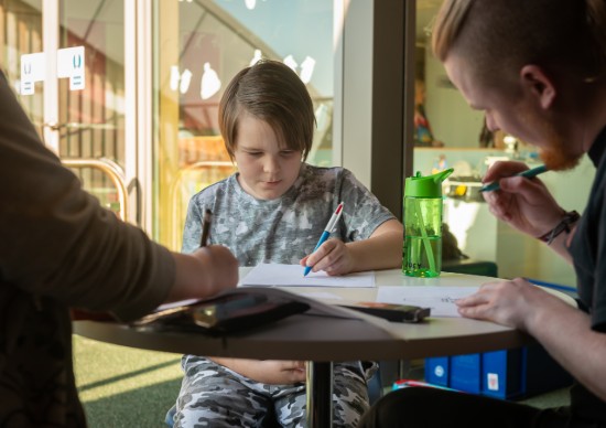 A young person sat at a table drawing, accompanied by two adults