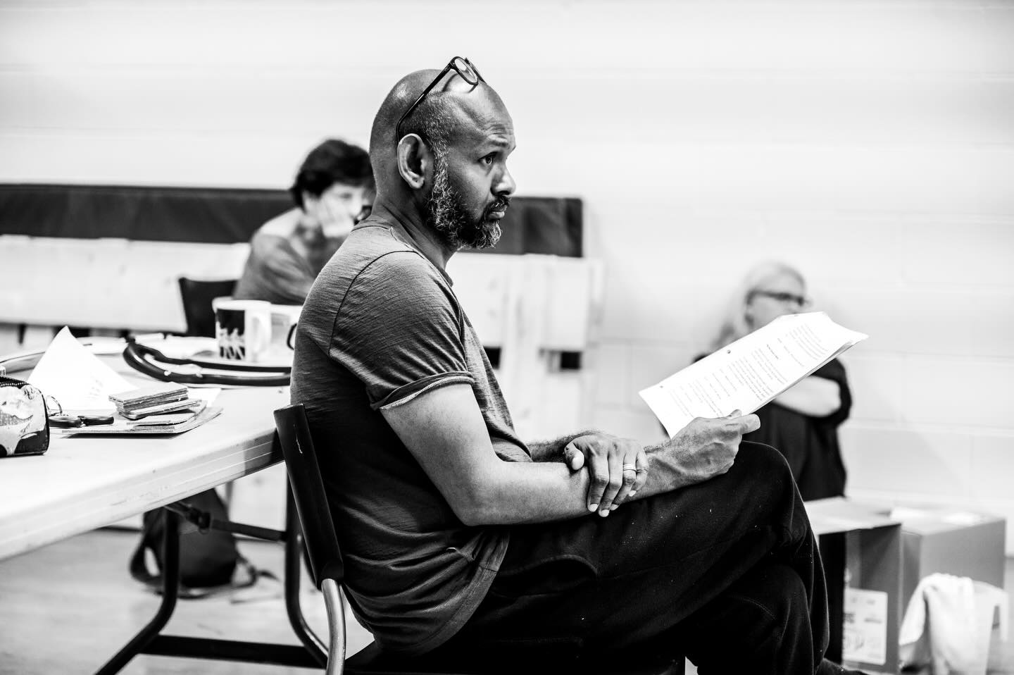 Ramesh is sat in a rehearsal room holding a script, glasses perched on his head. The image is in black and white.