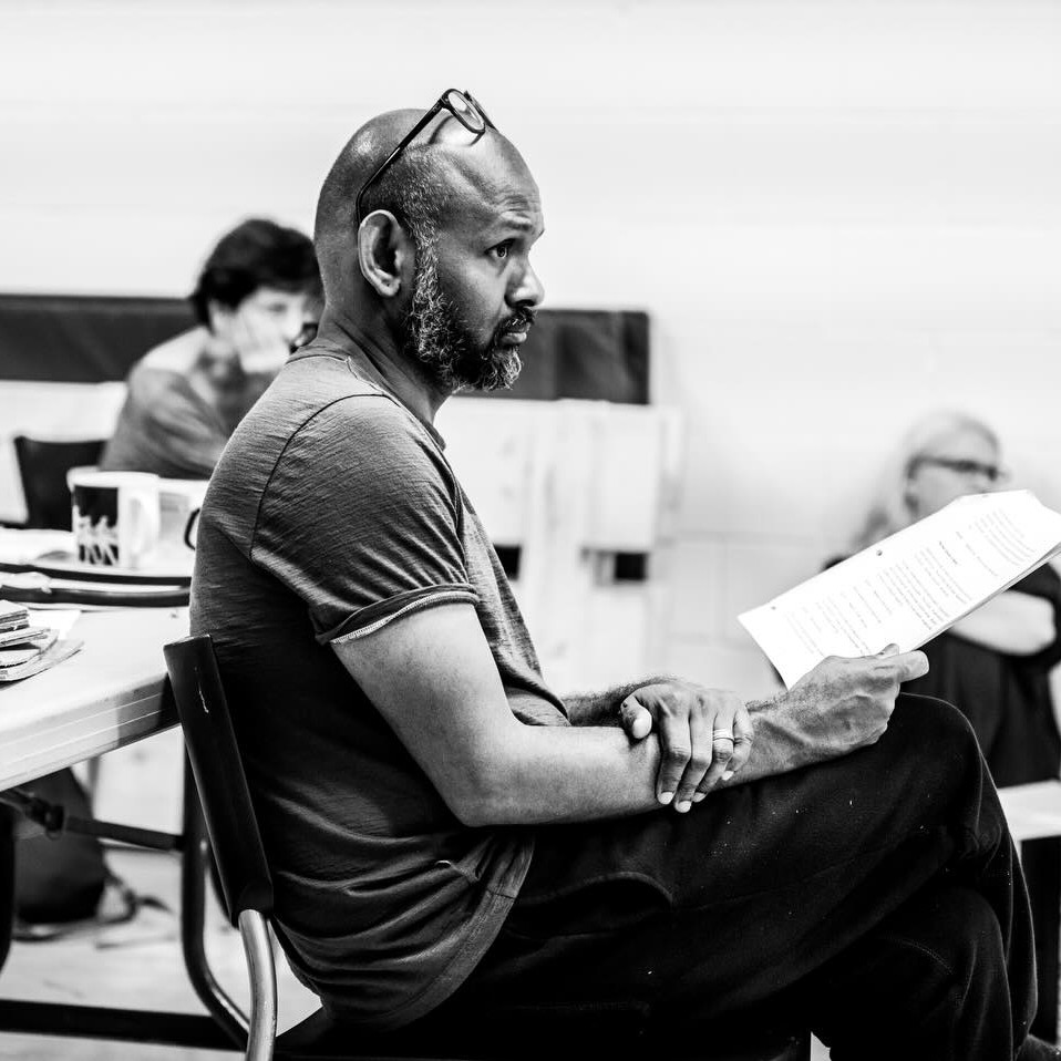 Ramesh is sat in a rehearsal room holding a script, glasses perched on his head. The image is in black and white.