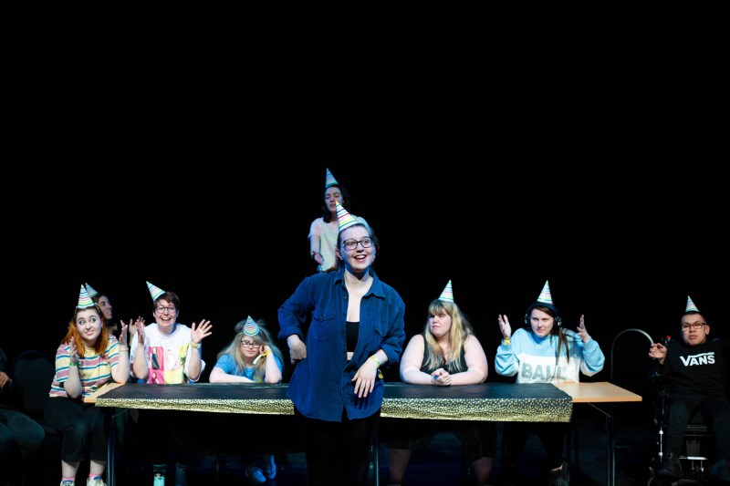 Young people onstage wearing party hats sat at a table. One of them is stood up and smiling in the centre of the image. 