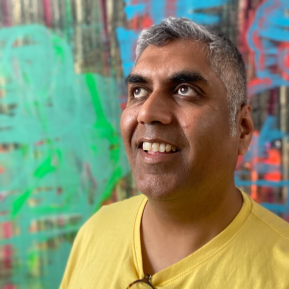 Headshot of Ashok Mistry, looking upwards and smiling. Ashok has grey short hair and is wearing a yellow t-shirt. Behind is colourful abstract art