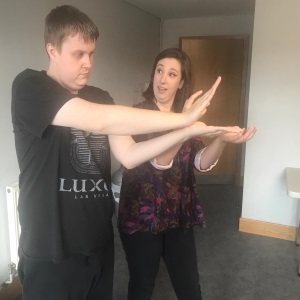 An image of a young man making shapes with his arms and hands. A young woman is next to him and looks like she is guiding him