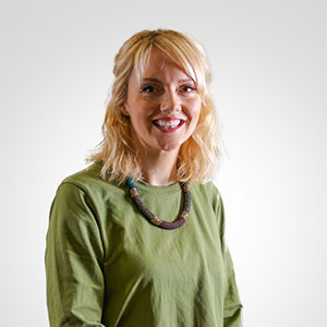 A photo of a woman with shoulder length blond hair wearing a green dress and a beaded necklace.