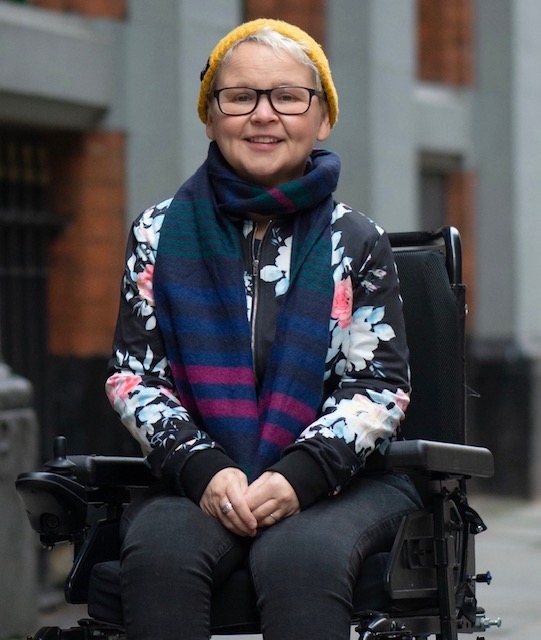 Nickie smiles into camera - she is outdoors wearing a woolly pullover and scarf.. Nickie is a wheelchair user.