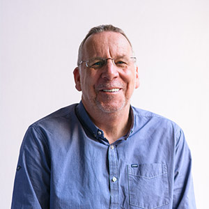 A photo of a man against a white background. He is wearing glasses and a blue shirt