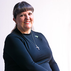 A photo of a woman against a white background. She is wearing a navy top and a necklace with a cross