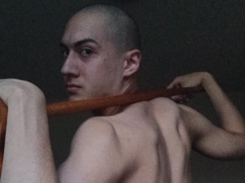 Chris Shapiro turns from behind to face camera resting arms across a stick which lies across their shoulders. Chris is topless with a shaved head.
