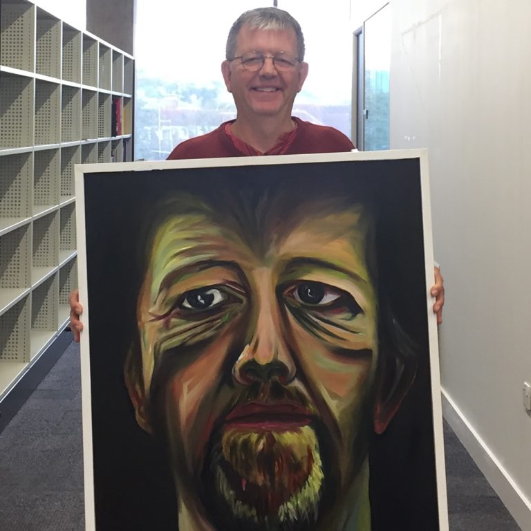 A white man stands holding a large painting of a face
